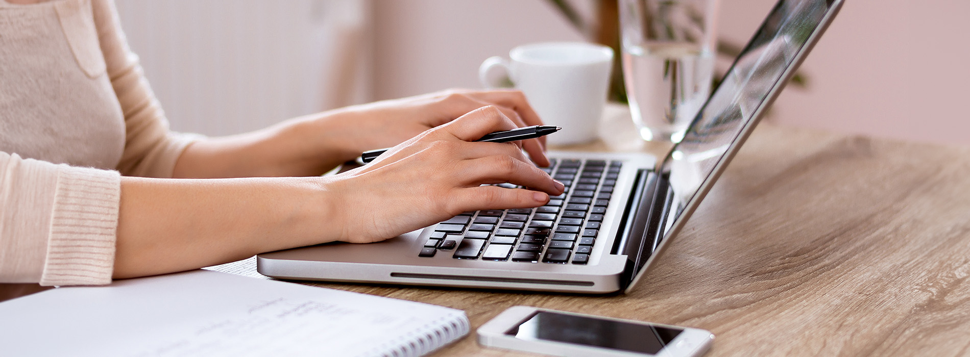 A person typing on a laptop at a desk with a notebook, pen, and smartphone nearby.