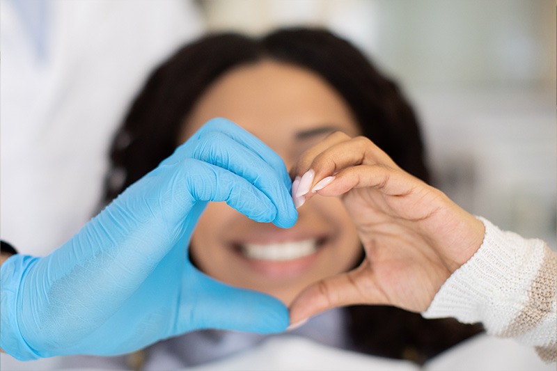 The image shows a person holding up a heart shape with their hands while wearing blue gloves, against a blurred background that includes medical equipment and personnel.