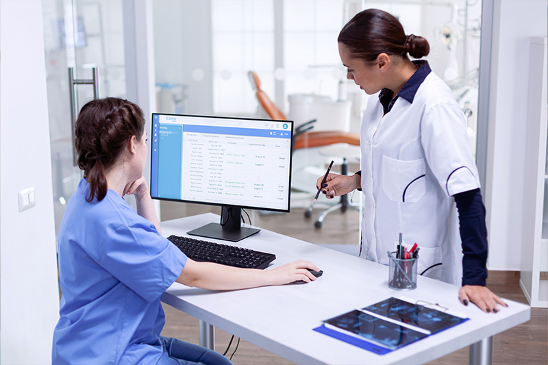 A professional healthcare setting with two individuals  a woman in scrubs working at a desk with a computer monitor displaying a webpage, and another woman standing behind her, both engaged in their tasks within a modern office environment.